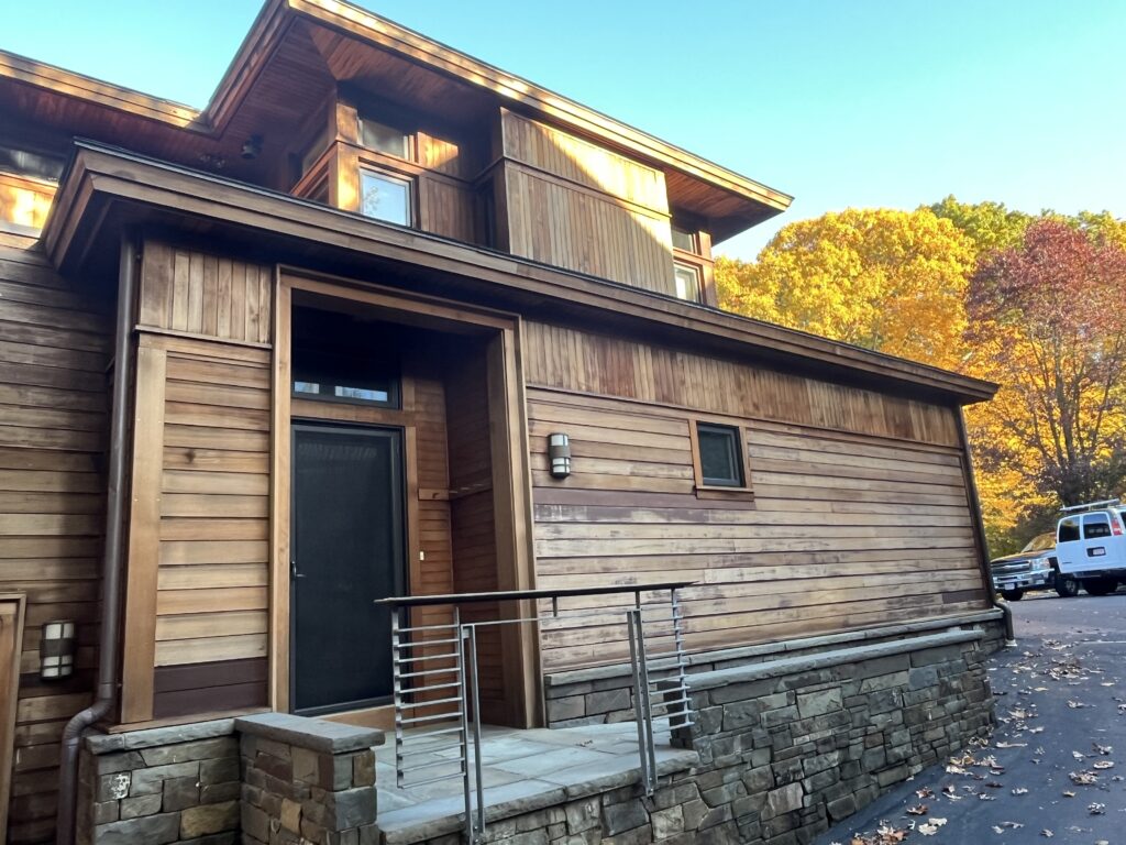Modern wooden house entrance featuring a stone base, glass door, and a metal railing. Vibrant autumn foliage adds color to the background.