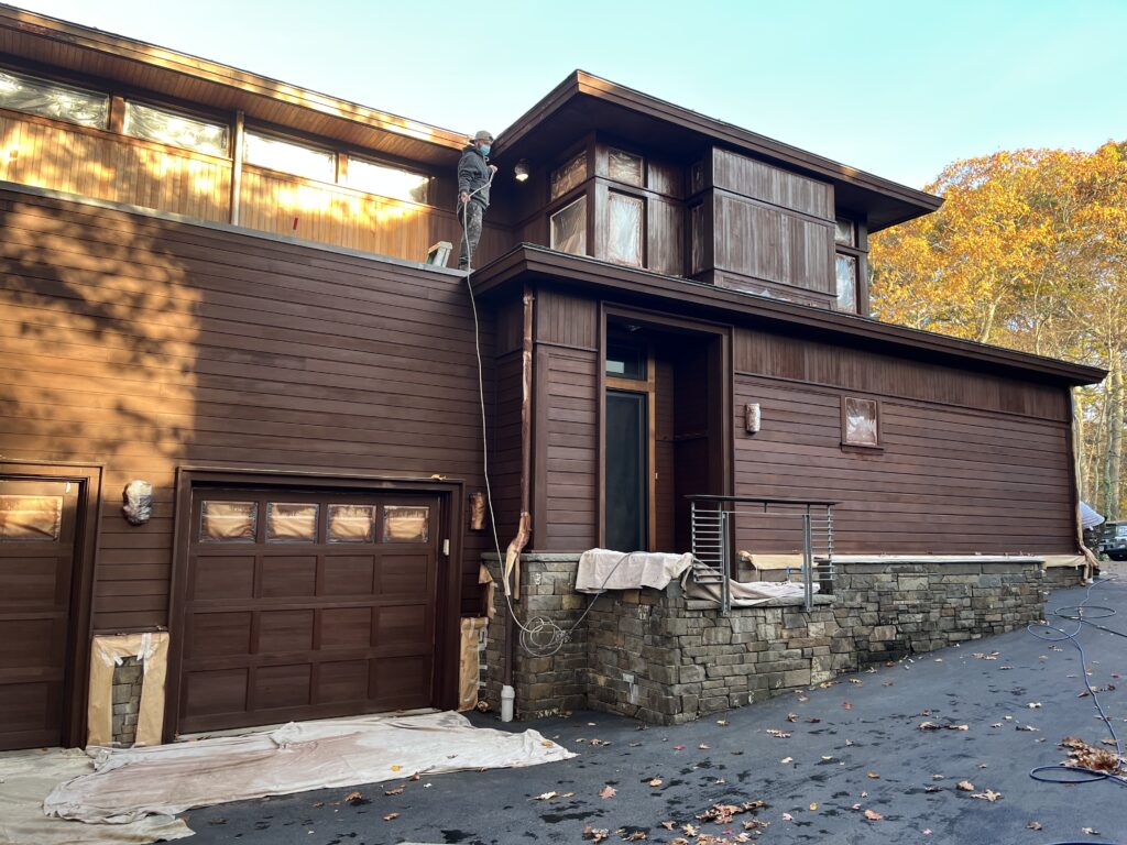 A worker paints the exterior of a modern wooden house, showcasing its rich brown color and stone accents, set against a backdrop of autumn foliage.