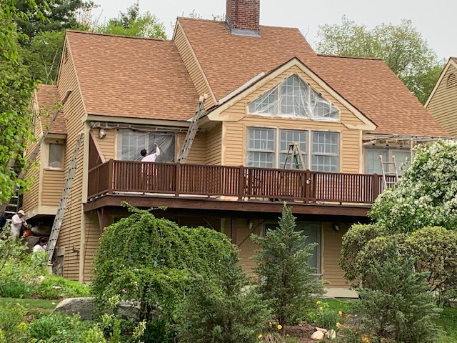 Two-story residential house with a wooden deck, large windows, and landscaped greenery surrounding the property.